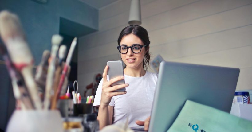 woman in white t shirt holding smartphone in front of laptop