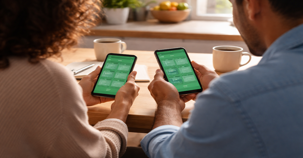 Two people sitting at a kitchen table each viewing matching budgeting app dashboards on their smartphones