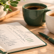 Two coffee mugs beside an open budget notebook on a kitchen table, representing the financial planning conversations couples have in their late 20s and 30s — housing decisions, shared budgets, and debt payoff strategy
