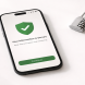 Smartphone displaying a security shield with green checkmark on a financial app beside a small combination lock on a white surface