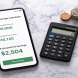 Smartphone showing a tax savings calculation screen with green numbers beside a calculator and stacked coins on white marble
