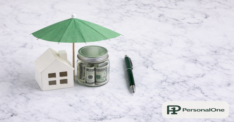 Small house figurine and glass jar of bills with a green paper umbrella placed over them representing financial protection on white marble — PersonalOne