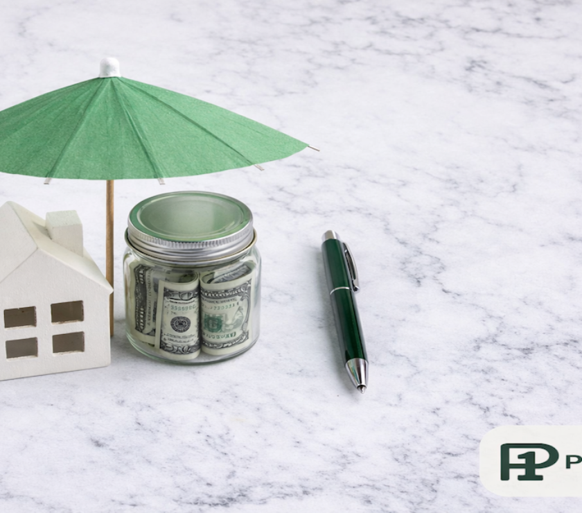 Small house figurine and glass jar of bills with a green paper umbrella placed over them representing financial protection on white marble — PersonalOne